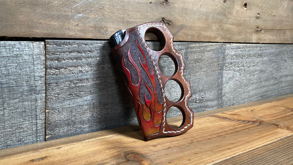 Knuckle duster brown leather lighter holder with flames leather tooled in red, gold and orange; brown polished leather with cream stitching detail. Holes for four fingers. Sitting on brown and grey rustic shelf.