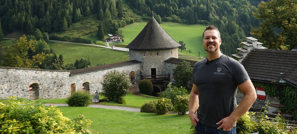 DIY patterns for Leather, Metal and Wood Fabrication. Levi smiling, hands in pockets of jeans, fitted drawbridge props t-shirt at Hohenwerfen Castle, large castle in background with turrets; rolling green lawn, shrubs and grey stone medieval castle wall f