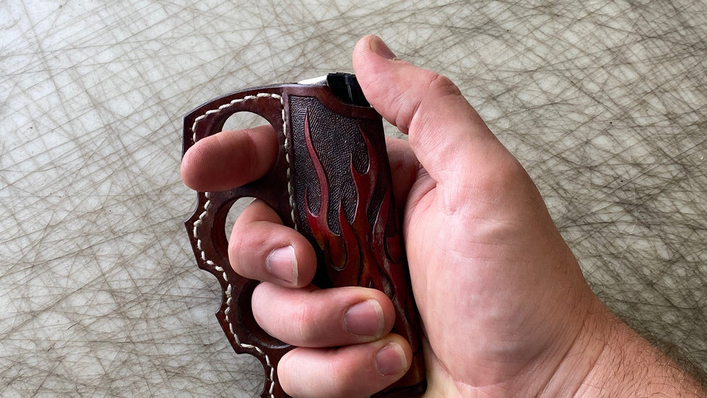 
                  
                    Hand holding knuckle duster brown leather lighter holder with flames leather tooled brown polished leather with cream stitching detail.
                  
                