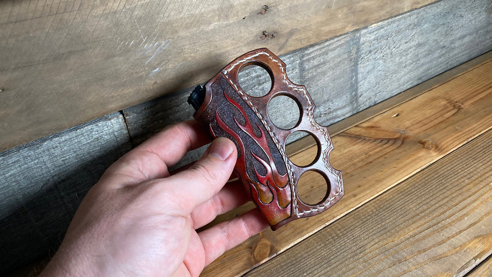 
                  
                    Hand holding knuckle duster brown leather lighter holder with flames leather tooled brown polished leather with cream stitching detail.
                  
                