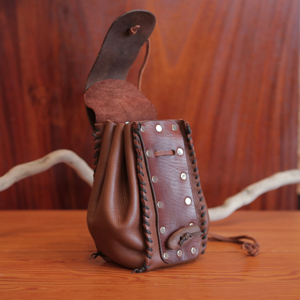 
                  
                    Side view of expandable brown leather handmade pouch, revealing second inner flap. Pouch has contrasting dark brown stitching, flat rivets, leather drawstring and rustic toggle to close at front.
                  
                