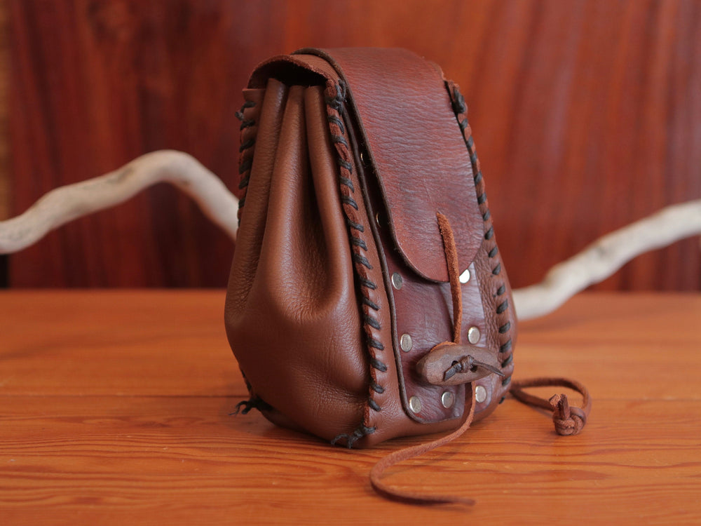 
                  
                    Side view of expandable brown leather handmade pouch with contrasting dark brown stitching, flat rivets, leather drawstring and rustic toggle to close at front.
                  
                